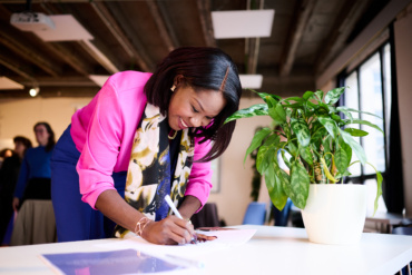 A black-skinned woman signs the petition at the table. The woman is smiling. She wears a light pink blazer, blue pants and a floral scarf.