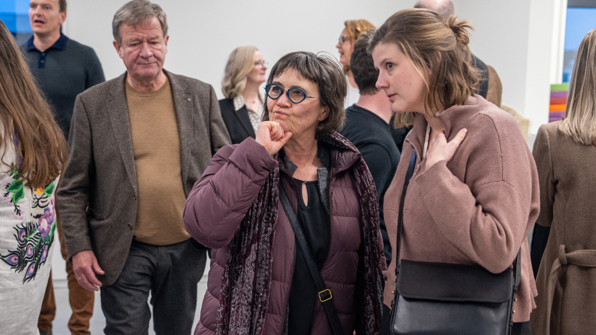 A picture on an older woman with grey hair and blue glasses. she is wearing a purple coat, purple scarf and a dark shirt. On her left, there is an older man wearing a grey jacket and a beige shirt. He is not looking at the woman. To the woman's right, there is a younger woman touching her chest. She is wearing a light pink sweater and a black bag. She is looking to the left and seem to be listening to the woman speaking.