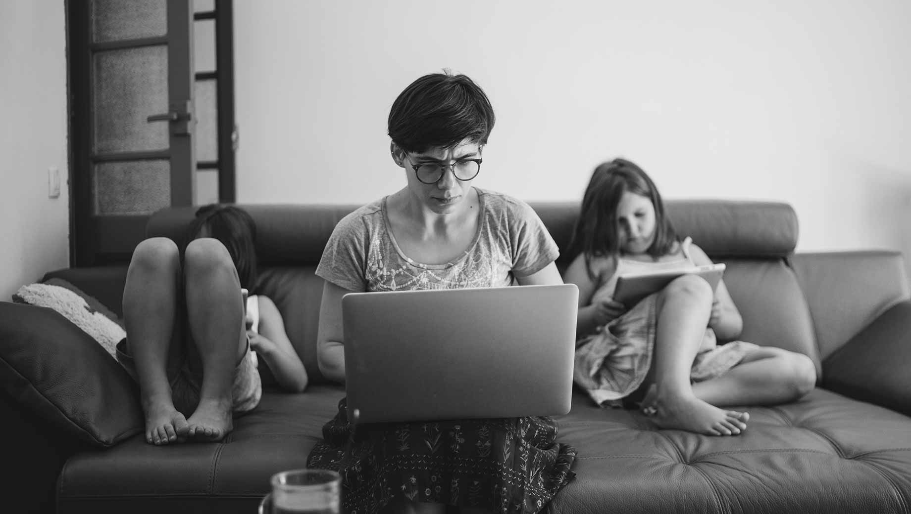 A black and white picture of a mother working on her computer. She is sitting in the middle of the sofa, with her two daughters by her side. The daughters are taking care of themselves, reading a book.