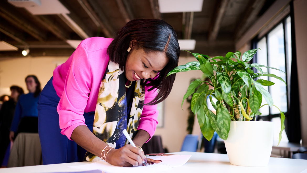 A black-skinned woman signs the petition at the table. The woman is smiling. She wears a light pink blazer, blue pants and a floral scarf.